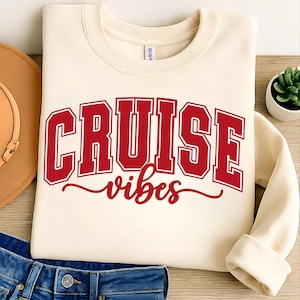 May include: Cream-colored sweatshirt with the words "CRUISE" and "vibes" in red block letters. The sweatshirt is folded, with a portion of blue jeans and a brown hat visible in the background. A small potted plant is also visible.