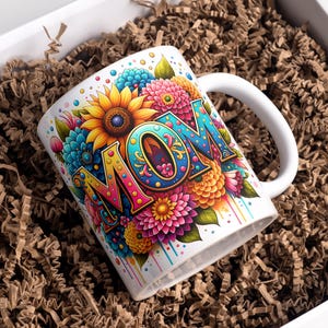 Könnte beinhalten: Eine weiße Keramiktasse mit einem bunten Blumenmuster und dem Wort "MOM" in großen, verzierten Buchstaben. Die Tasse befindet sich in einer Schachtel, die mit braunem Packpapier gefüllt ist.