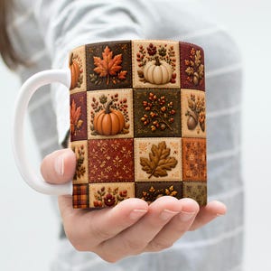May include: A ceramic mug with a white handle featuring a patchwork design of autumn-themed squares. The pattern includes illustrations of orange pumpkins, brown maple leaves, acorns, and berries in shades of brown, tan, and deep red.