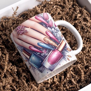 Op de afbeelding: Witte keramische mok met een nail art design. Het ontwerp toont een hand met lange, gelakte nagels in roze, blauw en goud, plus nagellakflesjes. De mok staat in een witte doos gevuld met bruin papier.