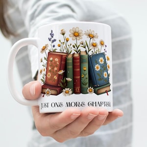 Op de afbeelding: Witte keramische mok met een ontwerp van boeken en bloemen, waaronder madeliefjes. De mok heeft de tekst "JUST ONE MORE CHAPTER". De mok wordt door een persoon vastgehouden.