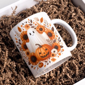 May include: White ceramic mug with a Halloween theme. The mug features a smiling ghost, a jack-o'-lantern, and sunflowers. The mug is in a white box with brown crinkle paper.