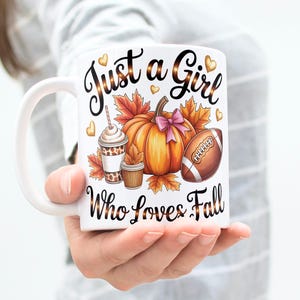 Puede incluir: Taza de cerámica blanca con un diseño de temática otoñal. La taza presenta una calabaza con un lazo rosa, un balón de fútbol americano y una taza de café. El texto en la taza dice "Just a Girl Who Loves Fall."