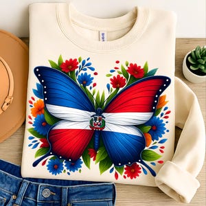 Könnte beinhalten: Cremefarbenes Sweatshirt mit einem Schmetterlingsdesign in den Farben der Flagge der Dominikanischen Republik: Blau, Rot und Weiß. Der Schmetterling ist von roten, blauen und orangen Blumen und grünen Blättern umgeben. Das Sweatshirt ist gefaltet.