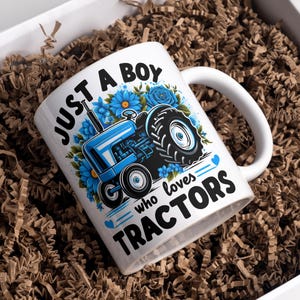 May include: White ceramic mug with a blue tractor design and the text "JUST A BOY who loves TRACTORS". The mug is decorated with blue flowers and hearts. The mug is in a white box with brown paper filler.