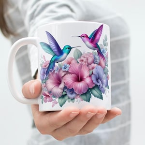 Könnte beinhalten: Weiße Keramiktasse mit zwei Kolibris in Blau-, Lila- und Rosatönen. Die Vögel sind vor einem Hintergrund aus rosa, lila und blauen Blumen abgebildet. Die Tasse hat einen gebogenen Griff.