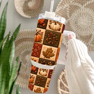 May include: A tall cylindrical tumbler with a handle and a clear straw, featuring a repeating grid pattern of autumn-themed squares. The design includes illustrations of orange pumpkins, brown oak leaves, and colorful fall foliage on a dark background.