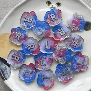 Puede incluir: Una colección de piezas de resina coloridas en forma de flor, cada una con una letra 'R' o 'L' incrustada. Las piezas son en tonos de azul, rosa y morado, con acentos de purpurina. Están dispuestas sobre un plato blanco con forma de gato.