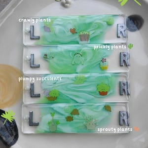 Puede incluir: Cuatro horquillas rectangulares transparentes con temática de plantas con las letras "L" y "R". Cada horquilla tiene un tema de planta diferente: plantas rastreras, plantas espinosas, suculentas regordetas y plantas brotantes. Las horquillas están sobre un plato blanco con un diseño de cara de gato.