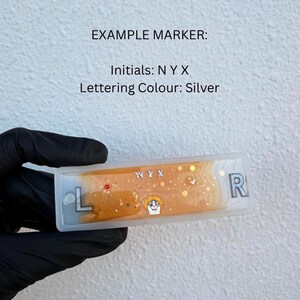 Può includere: Un pennarello rettangolare con le iniziali "NYX" in lettere argentate. Il pennarello ha un interno dorato e arancione con elementi decorativi. Le lettere "L" e "R" sono su entrambe le estremità del pennarello. È visibile anche il testo "EXAMPLE MARKER: Initials: N Y X Lettering Colour: Silver".