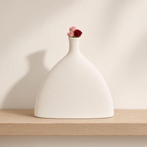 Op de afbeelding: Een matwitte keramische vaas met een brede, driehoekige basis en een smalle hals. De vaas heeft een glad, effen oppervlak en bevat drie kleine, delicate rozenbloesems in tinten lichtroze en donkerrood bij de opening aan de bovenzijde.