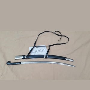 May include: A curved steel sword with a black handle and a matching black scabbard featuring silver-toned metal accents. A black leather shoulder strap is attached to the scabbard. A small white paper tag with the handwritten text swordrep is included.
