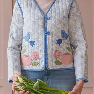 Peut inclure: Veste matelassée bleu clair avec un motif floral et d'oiseaux. La veste a un col en V, des manches longues et une fermeture à boutons. La veste est ornée de fleurs roses appliquées et d'oiseaux bleus. La personne porte un jean bleu.