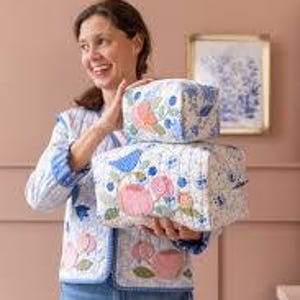 Puede incluir: Dos neceseres acolchados blancos con bordados florales y de pájaros están apilados. Los sostiene una persona que lleva una chaqueta acolchada a juego y vaqueros azules. Las bolsas presentan diseños rosas, azules y verdes.