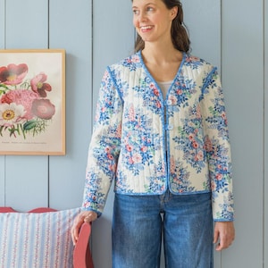 Puede incluir: Chaqueta acolchada con estampado floral en tonos azules, rosas y blancos, con ribete azul y cuello en V. La persona lleva vaqueros azules. Se ve un estampado floral enmarcado y un cojín a rayas. Diseño clásico.