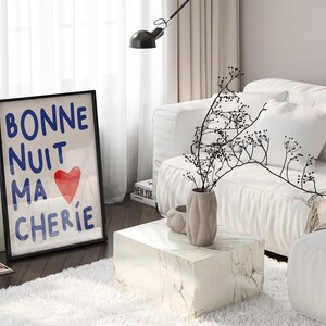 May include: A black framed print with the text "Bonne Nuit Ma Cherie" in blue lettering and a red heart. The print is on a white marble side table in a living room with a white couch and a vase with dried branches.