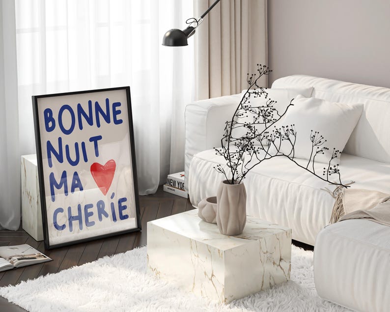 May include: A black framed print with the text "Bonne Nuit Ma Cherie" in blue lettering and a red heart. The print is on a white marble side table in a living room with a white couch and a vase with dried branches.