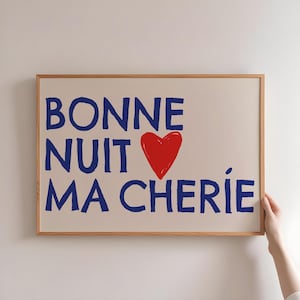 May include: A framed art print with the words "BONNE NUIT MA CHERIE" in blue, a red heart, and a light beige background. The frame is made of light brown wood. The print is held by a person's hand.
