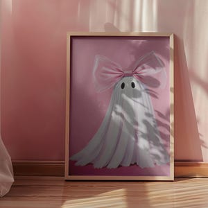 May include: A framed print featuring a whimsical ghost design. The ghost is white with a large pink bow and two black dot eyes, set against a pink background. The frame is a light wood color. The artwork is displayed on a pink wall.