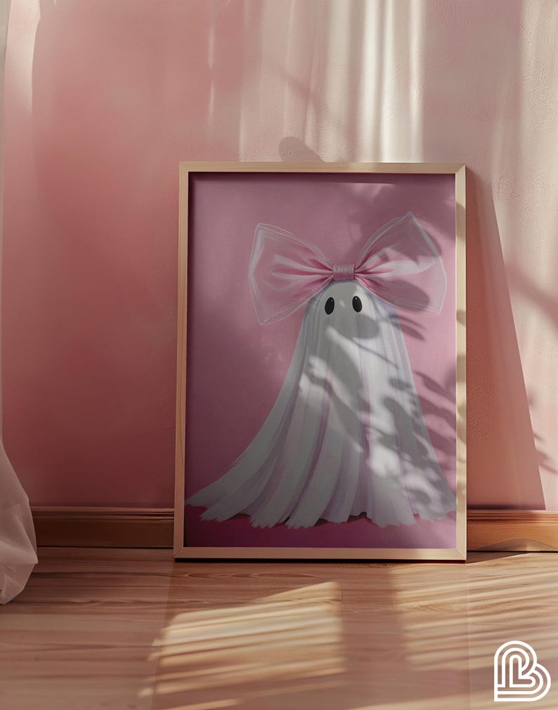 May include: A framed print featuring a whimsical ghost design. The ghost is white with a large pink bow and two black dot eyes, set against a pink background. The frame is a light wood color. The artwork is displayed on a pink wall.