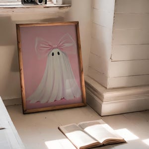 May include: A framed artwork featuring a ghost with a pink bow and a pink background. The ghost has two black eyes. A vintage camera sits on a windowsill. An open book rests on the floor, illuminated by sunlight.