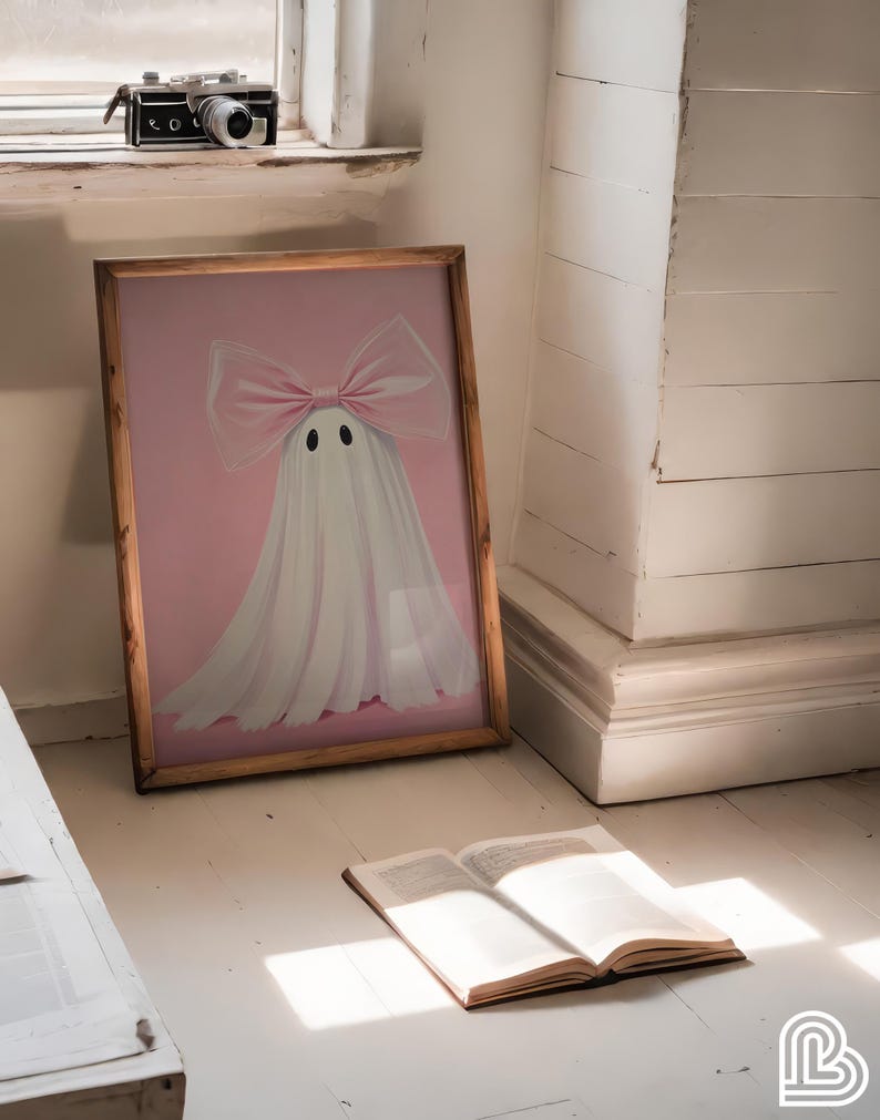 May include: A framed artwork featuring a ghost with a pink bow and a pink background. The ghost has two black eyes. A vintage camera sits on a windowsill. An open book rests on the floor, illuminated by sunlight.