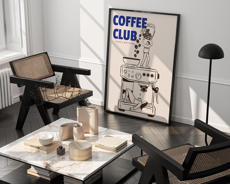 May include: A framed wall art print featuring the text COFFEE CLUB every morning in blue and red. The illustration depicts a person standing on a coffee machine pouring beans from a jar, while another person lies underneath the machine.
