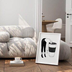 May include: A framed black and white illustration depicts a person leaning into a large coffee cup. The artwork is set against a neutral-toned room with a modern, rounded sofa and a cup of coffee on a stack of books.