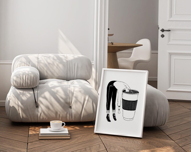 May include: A framed black and white illustration depicts a person leaning into a large coffee cup. The artwork is set against a neutral-toned room with a modern, rounded sofa and a cup of coffee on a stack of books.