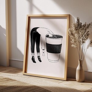 May include: A framed art print featuring a black and white illustration of a person leaning over a large coffee cup. The person is wearing black pants, a white sweater, and black boots. The artwork is displayed in a light wood frame.