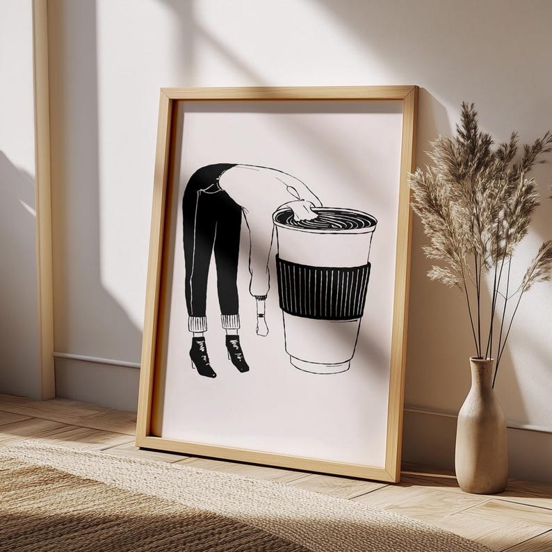 May include: A framed art print featuring a black and white illustration of a person leaning over a large coffee cup. The person is wearing black pants, a white sweater, and black boots. The artwork is displayed in a light wood frame.