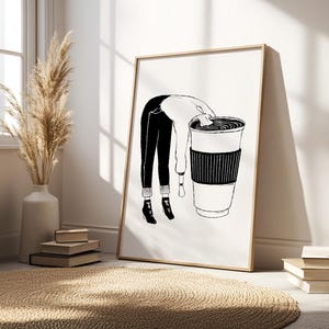 May include: Black and white art print of a person leaning into a large coffee cup. The person is wearing black pants, a white sweater, and black boots. The coffee cup has a black sleeve. The art print is in a wooden frame.