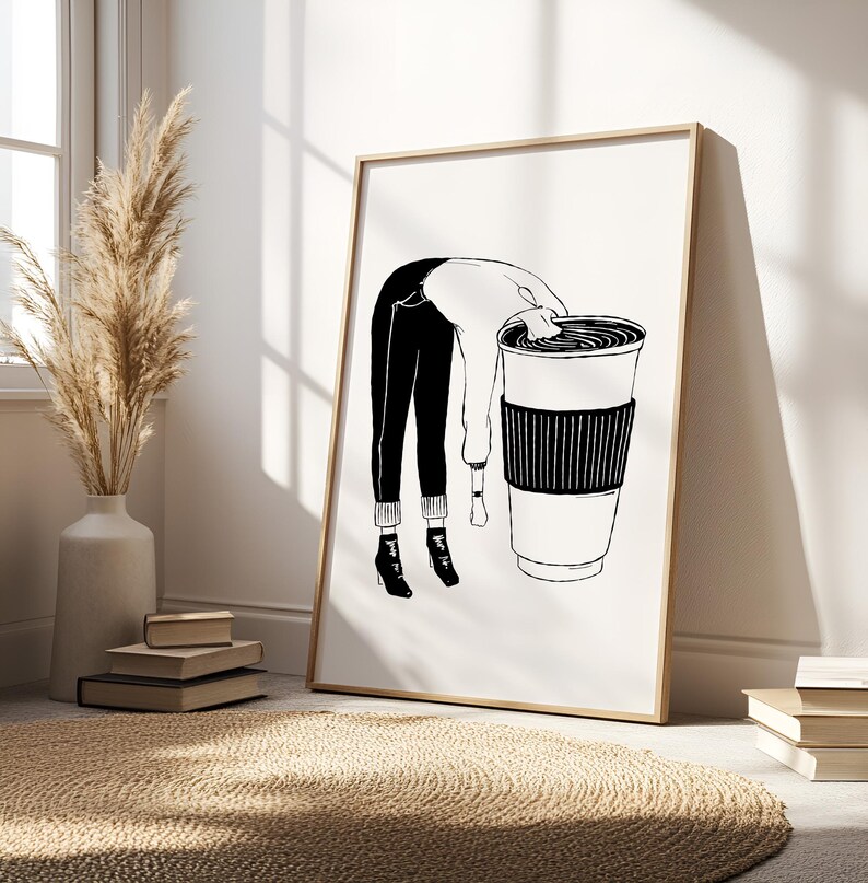May include: Black and white art print of a person leaning into a large coffee cup. The person is wearing black pants, a white sweater, and black boots. The coffee cup has a black sleeve. The art print is in a wooden frame.