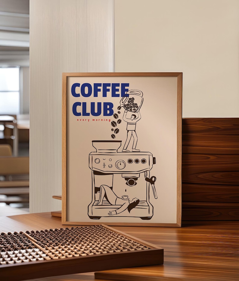 May include: A framed poster with a light beige background featuring the blue text COFFEE CLUB and the red text every morning. The illustration shows a person pouring coffee beans into a machine while another person lies underneath it.