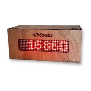 Może przedstawiać: Prostokątny drewniany wyświetlacz cyfrowy z czerwonym napisem "1660". Wyświetlacz znajduje się w jasnobrązowej drewnianej obudowie z logo "Sweetco" i tekstem "sweetcounter".