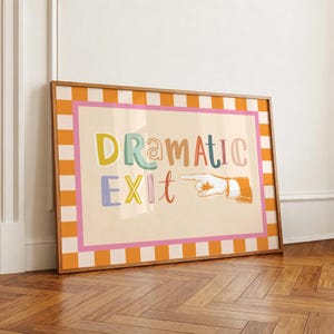 May include: A framed rectangular wall art piece featuring a cream-colored background with the text "DRAMATIC EXIT" in multicolored, playful lettering. To the right of the text is a line-art illustration of a hand pointing to the right.
