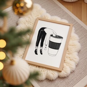 May include: A framed black and white illustration depicts a person leaning into a large coffee cup. The person's legs and torso are visible, with the head obscured. The cup has a ribbed sleeve. The artwork is set on a fluffy, white rug.
