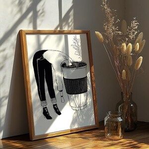May include: A framed black and white illustration depicts a person bending over a large coffee cup. The person is wearing black pants, a white sweater, and black boots. The cup has a black sleeve. A vase of dried flowers and a glass jar are in the background.