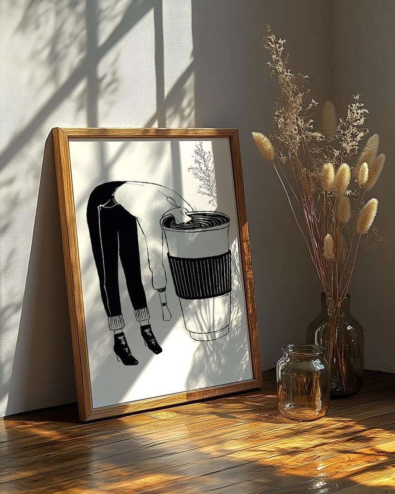 May include: A framed black and white illustration depicts a person bending over a large coffee cup. The person is wearing black pants, a white sweater, and black boots. The cup has a black sleeve. A vase of dried flowers and a glass jar are in the background.