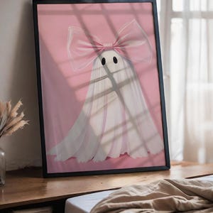 May include: Framed art print featuring a whimsical ghost design. The ghost is white with a large pink bow and black eyes, set against a pink background. The artwork is in a black frame, suitable for home decor.