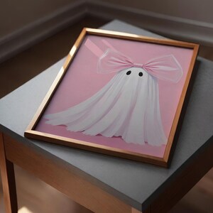 May include: A framed artwork featuring a whimsical ghost design. The ghost is depicted with a white sheet and a large pink bow. The background is a soft pink, and the frame is a warm gold color. The artwork is displayed on a gray table.