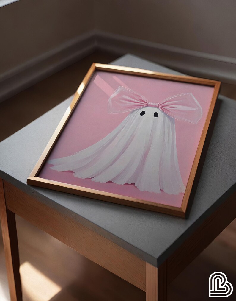 May include: A framed artwork featuring a whimsical ghost design. The ghost is depicted with a white sheet and a large pink bow. The background is a soft pink, and the frame is a warm gold color. The artwork is displayed on a gray table.