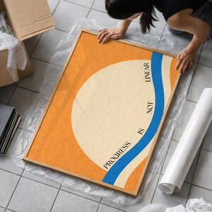 May include: Framed art print with an abstract design. The artwork features a large off-white circle against an orange background, with a blue wave and the text "PROGRESS IS NOT LINEAR". The print is in a wooden frame.