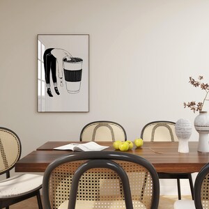 May include: Black and white art print featuring a person leaning into a large coffee cup. The person is wearing black pants and a white shirt. The coffee cup has a black sleeve. The art print is displayed on a wall.