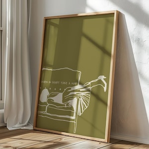 May include: A framed art print with a minimalist white illustration on an olive green backdrop. The artwork shows a person resting on a sofa, accompanied by the text "WHEN IN DOUBT TAKE A NAP". The frame is crafted from light-toned wood.