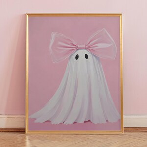 May include: A framed art print featuring a whimsical ghost illustration. The ghost is draped in white fabric and adorned with a large pink bow. The background is a soft pink, and the frame is gold.