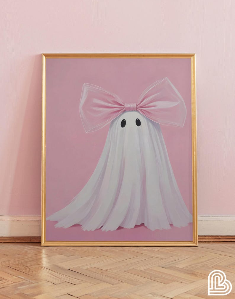 May include: A framed art print featuring a whimsical ghost illustration. The ghost is draped in white fabric and adorned with a large pink bow. The background is a soft pink, and the frame is gold.