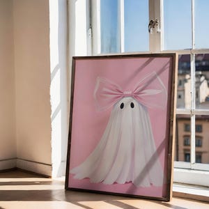 May include: A framed artwork featuring a whimsical ghost design. The ghost is depicted with a large pink bow and a flowing white sheet against a pink background. The artwork is in a wooden frame and is leaning against a window.