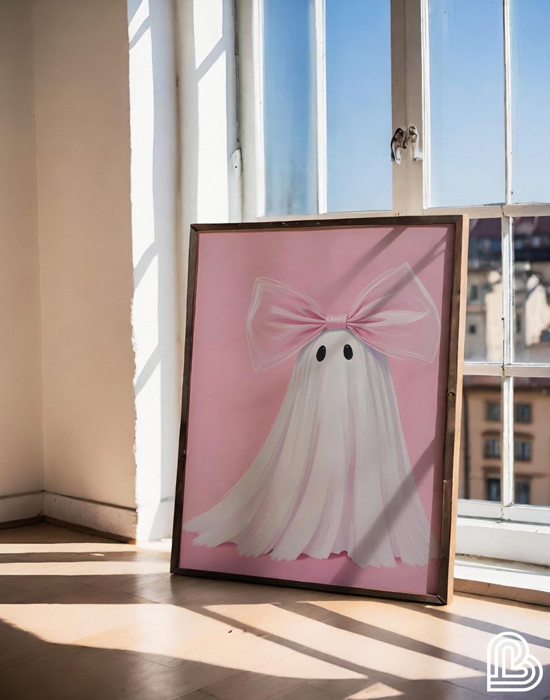 May include: A framed artwork featuring a whimsical ghost design. The ghost is depicted with a large pink bow and a flowing white sheet against a pink background. The artwork is in a wooden frame and is leaning against a window.