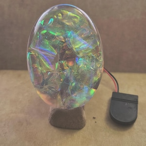 May include: An oval-shaped, clear resin sculpture containing iridescent, crinkled foil that reflects green, blue, and gold light. The sculpture is mounted on a small, brown wooden base and is connected to a black plastic battery pack by two wires.